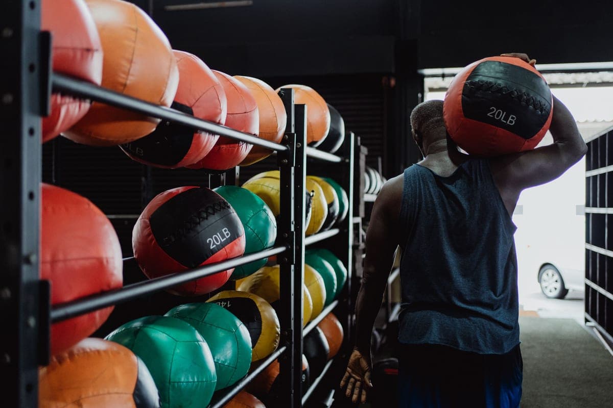 A man carrying a weighted ball past a rack of medicine balls in a gym